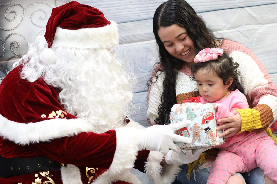 Isela Molina sonríe mientras su hija Yetzali Ayala, de 1 año, recibe un regalo de Santa durante el evento de Navidad de la Familia Migrante 2023 que se llevó a cabo en la biblioteca de la Firebaugh High School el 13 de diciembre.