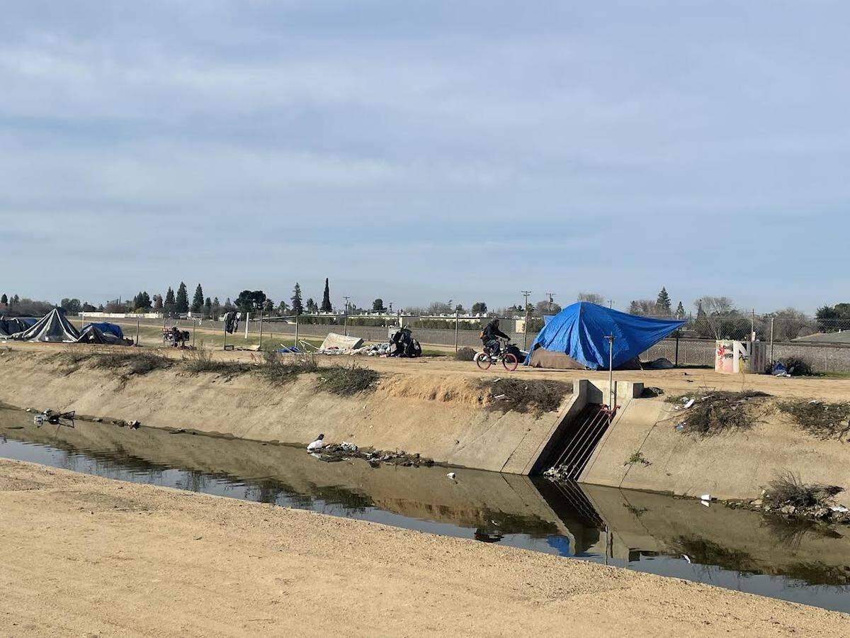 A bicyclist makes his way past an unhoused encampment on a Fresno Irrigation District canal near Palm and Dakota avenues on Friday, Feb. 23, 2024.