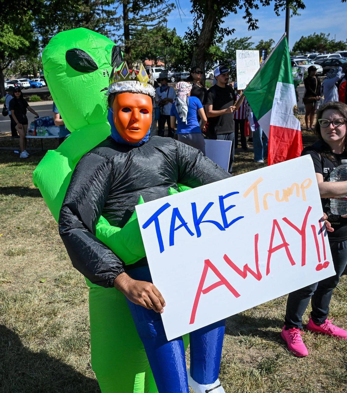 A protester dressed as an alien holding a crowned man with an orange face and an anti-Trump sign attends a rally at Cary Park near Fashion Fair Mall in Fresno before the start of a “No Kings” protest against President Donald Trump’s policies on Saturday, June 14, 2025.
