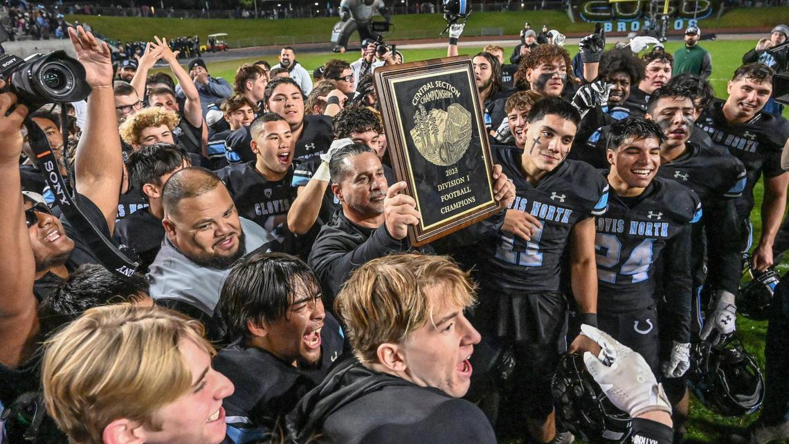 Clovis North head football coach Mike Jacot, center, holds up the 2023 Central Section Division 1 championship plaque while surrounded by players and staff after defeating Central 24-24 at Veterans Memorial Stadium in Clovis on Friday, Nov. 24, 2023.