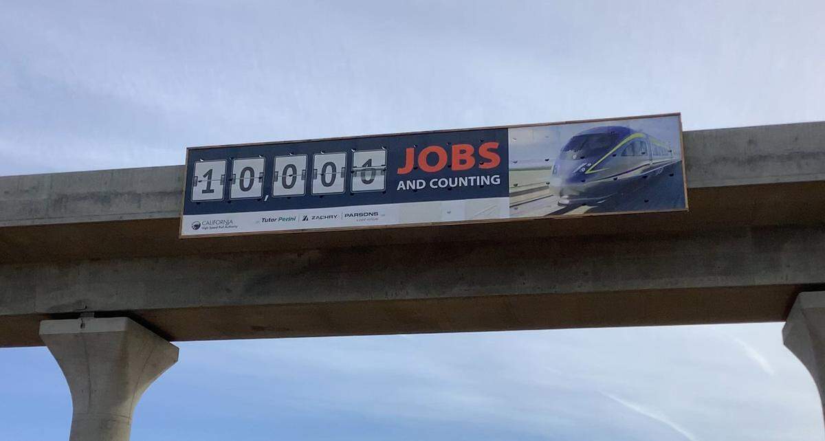 A sign affixed to the structure of a high-speed rail viaduct over Cedar Avenue near Highway 99 in south Fresno on Tuesday, Feb. 14, 2023, boasts the California High-Speed Rail Authority reaching a milestone of creating 10,000 jobs for construction of the project in the San Joaquin Valley.