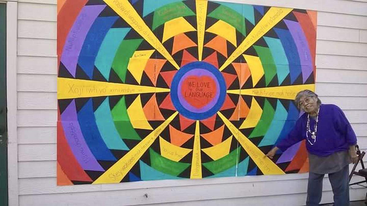 Marie Wilcox beside a mural promoting the preservation of indigenous languages.