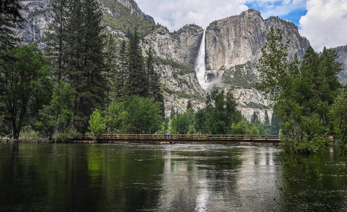 The bridge at the Swinging Bridge picnic area in Yosemite Valley sits just above the water level of the Merced River as Upper Yosemite Falls flows in the background on Tuesday, June 13, 2023.
