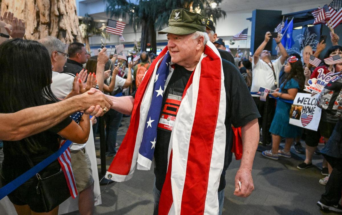 Veteran Lance Mackay of Madera walks through a giant crowd of welcomers shaking hands while wearing an American flag draped around his neck after arriving back at Fresno Yosemite International Airport along with 62 other veterans from the Central Valley Honor Flight on Wednesday, May 17, 2023.