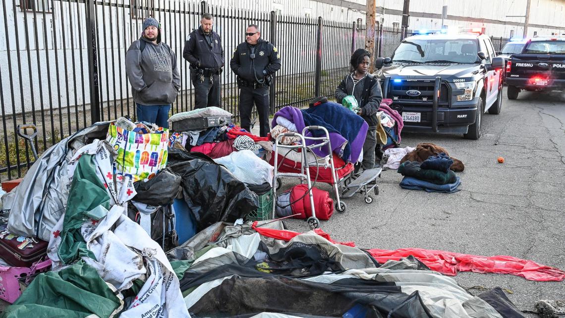 Fresno police officers stand near a pair of homeless people with their belongings pulled to the side of the street as Fresno’s Homeless Assistance Response Team (HART) begins a homeless camp cleanup operation behind several shelters on Parkway Drive in Fresno on Wednesday, Feb. 1, 2023