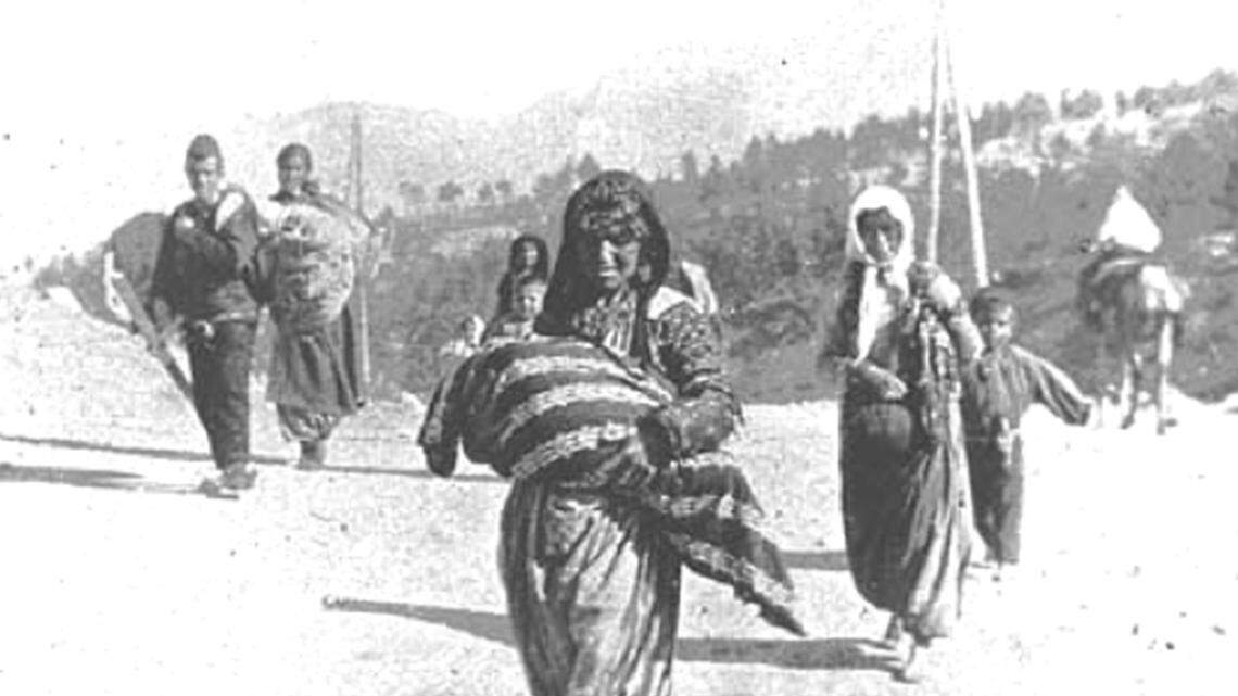In 1915, Armenian deportees _ women, children and elderly men _ during the genocide. The woman in the foreground is carrying a child in her arms, shielding it from the sun with a shawl; a man on left is carrying bedding; no other belongings or food noticeable are among effects being carried. All are walking in the sun on an unpaved road with no means of shelter from the elements. The location is Syria under the Ottoman Empire.