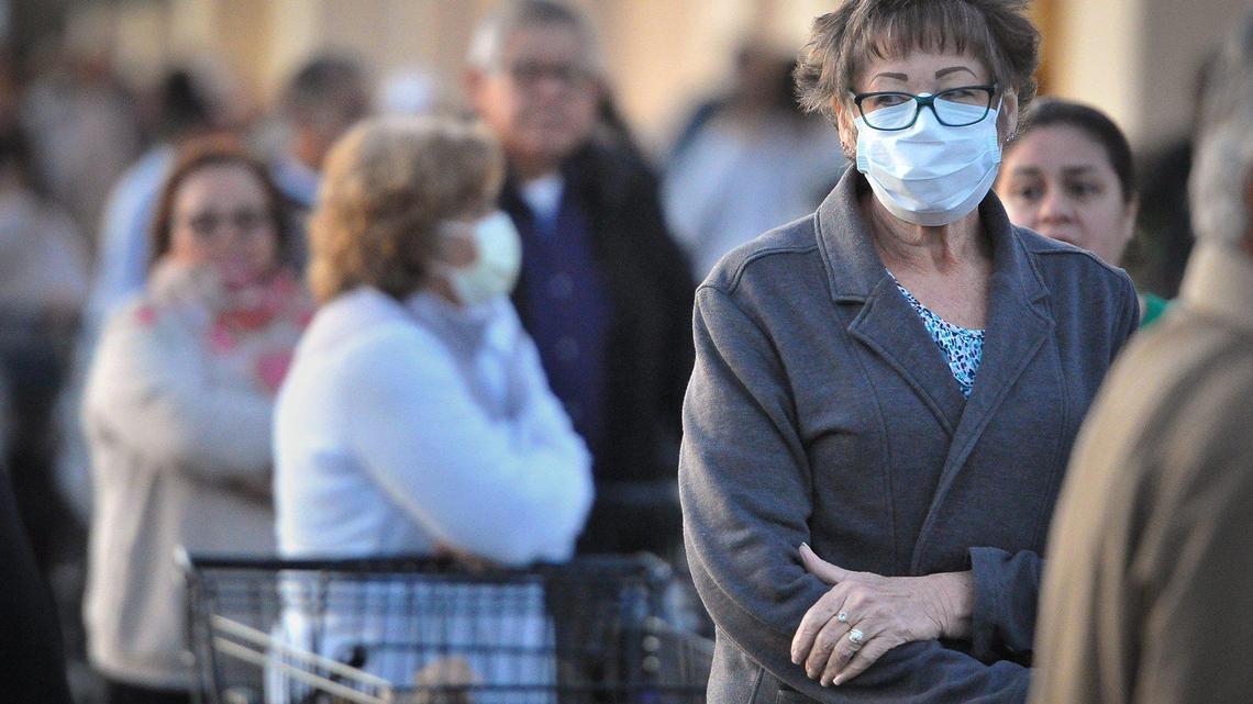 A woman in a protective mask waits in line at the Vallarta Supermarkets near Clinton Avenue and Highway 99 in Fresno with hundreds of others early Wednesday, March 18, 2020, amidst the coronavirus crisis.