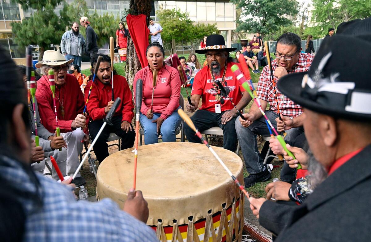 A drum circle plays and sings with a red dress hanging from a tree in the background as local families and relatives gathered at Fresno State’s Peace Garden to mark Missing and Murdered Indigenous Women and Girls’ Day, Friday, May 5, 2023 in Fresno.