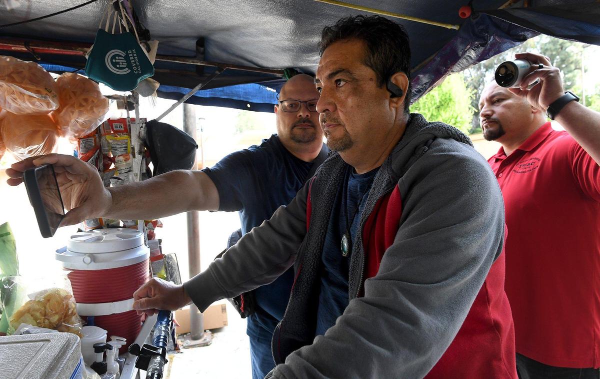 Pablo Villagrana, left, holding a security camera, and Joseph Vasquez, back left, demonstrate the use of the system designed to keep Fresno street vendors, such as Miguel Ruiz, center, safe as they work in Fresno, during a press conference at the Mosqueda Center, Wednesday March 30, 2022. The video feed, as shown on a mobile phone will be feed back to the Cloud system.