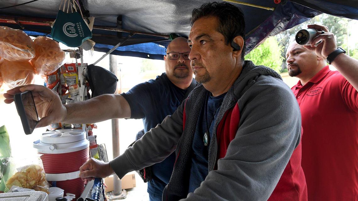 Pablo Villagrana, left, holding a security camera, and Joseph Vasquez, back left, demonstrate the use of the system designed to keep Fresno street vendors, such as Miguel Ruiz, center, safe as they work in Fresno, during a March 2022 press conference at the Mosqueda Center in southeast Fresno. The video feed, shown on a mobile phone will be feed back to a cloud storage system.