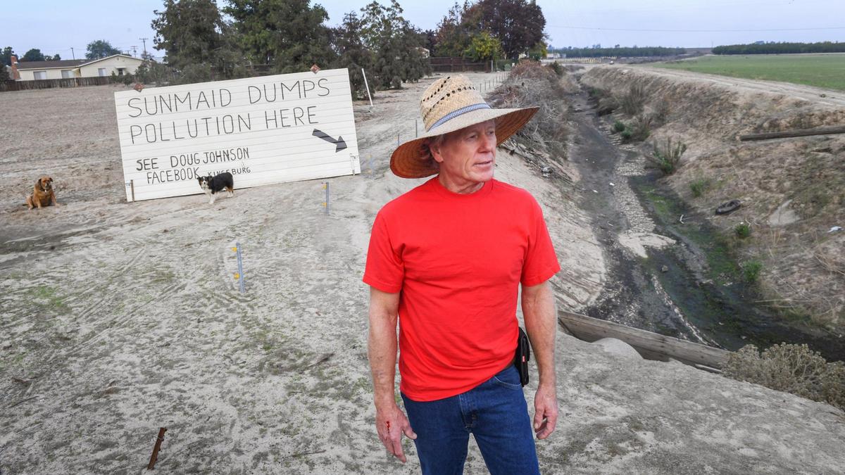 Kingsburg resident Doug Johnson, who lives next door to the Sun-Maid Raisin factory along Highway 99, stands near to a giant sign he made with a garage door and a bordering drainage canal on Wednesday, Nov. 11, 2021. Johnson says the factory dumped a massive amount of waste water into the canal that filled it with a black sludge.