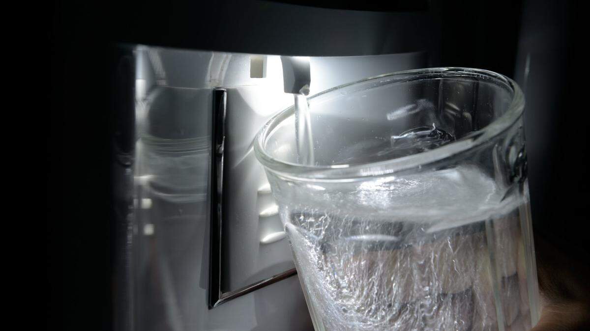“A glass of water being held up to a refrigerator’s water dispenser, the glass illuminated by the fridge’s light.”