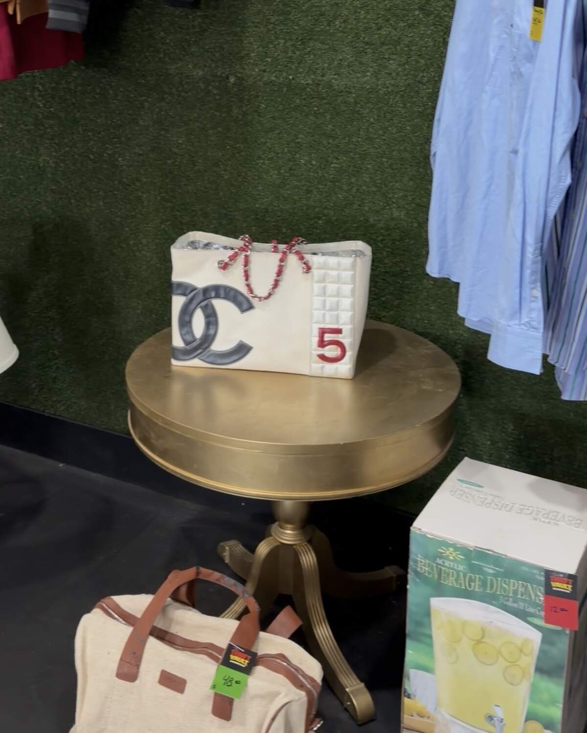A Chanel bag is displayed on a stand in front of a grass wall.