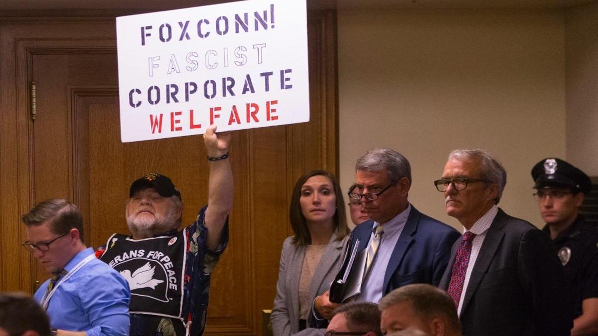 A lone protester holds up a sign as the Wisconsin Assembly Committee on Jobs and Economy meets about the incentive deal for Taiwan-based Foxconn Technology Group, Thursday, Aug. 3, 2017, at the state Capitol in Madison, Wis. The company plans to construct the first liquid crystal display panel factory outside of Asia in southeast Wisconsin. The deal requires the state to approve $3 billion in tax breaks tied to Foxconn hiring and spending as promised.