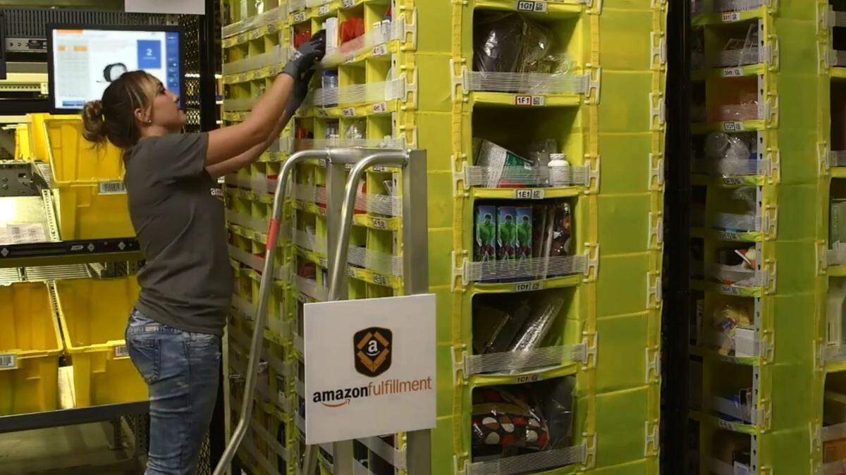An Amazon warehouse worker picks merchandise from a bin as she assembles a customer order for eventual shipping in this image from a company video inside one of its online-order fulfillment centers. Amazon opened its 855-000-square-foot e-commerce order-filling center in June with plans to hire about 1,500 employees.