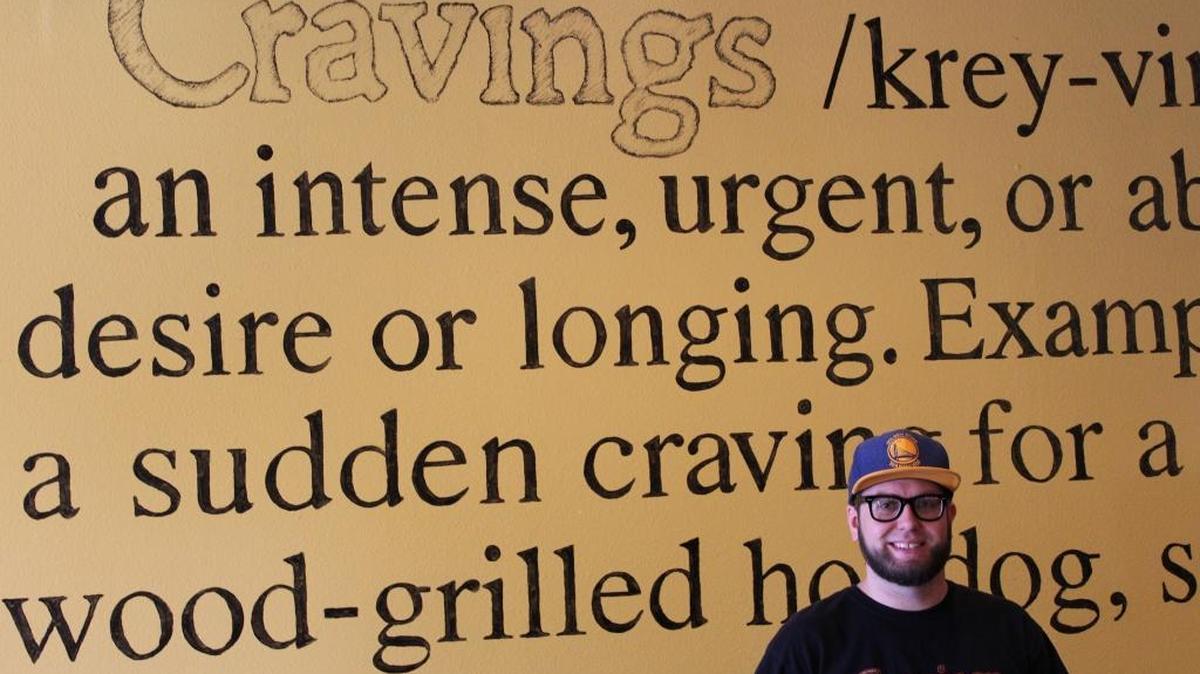 Cravings co-owner Grant Mohler stands next to a wall customized with a definition of what else? — cravings.