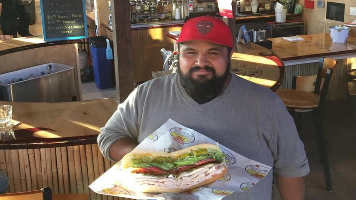 Sammy Franco displays a Surfin’ Bird, one of the gourmet sandwiches available at his Beach Hut Deli. The tiki hut thatch roof over the deli’s bar is in the background.