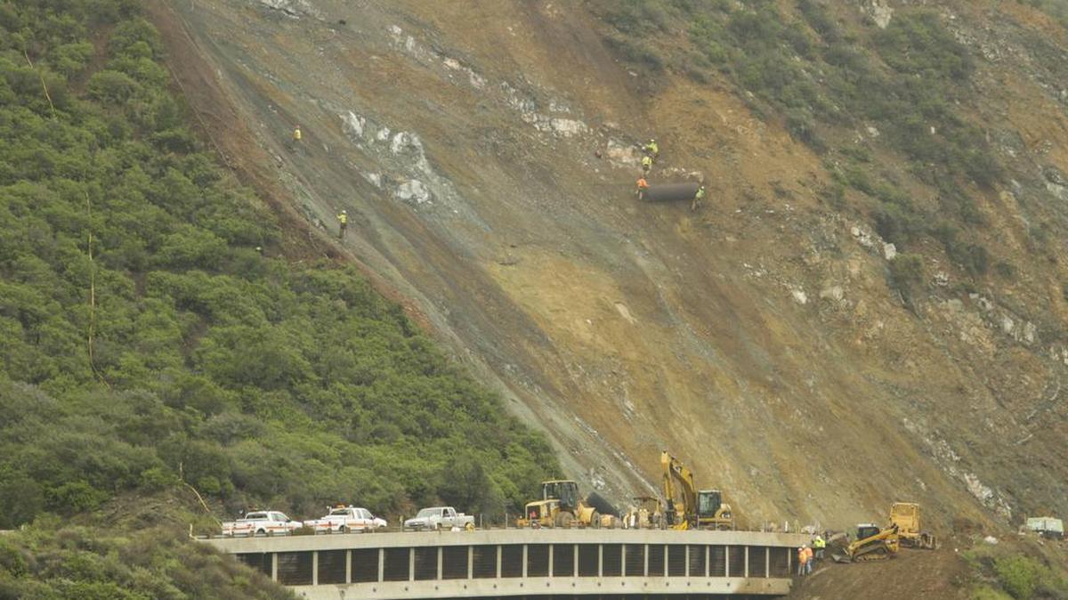 Crews unroll 75 foot long sections of steel mesh. The mesh allows rocks to fall in a controlled low energy fashion to the roadway.