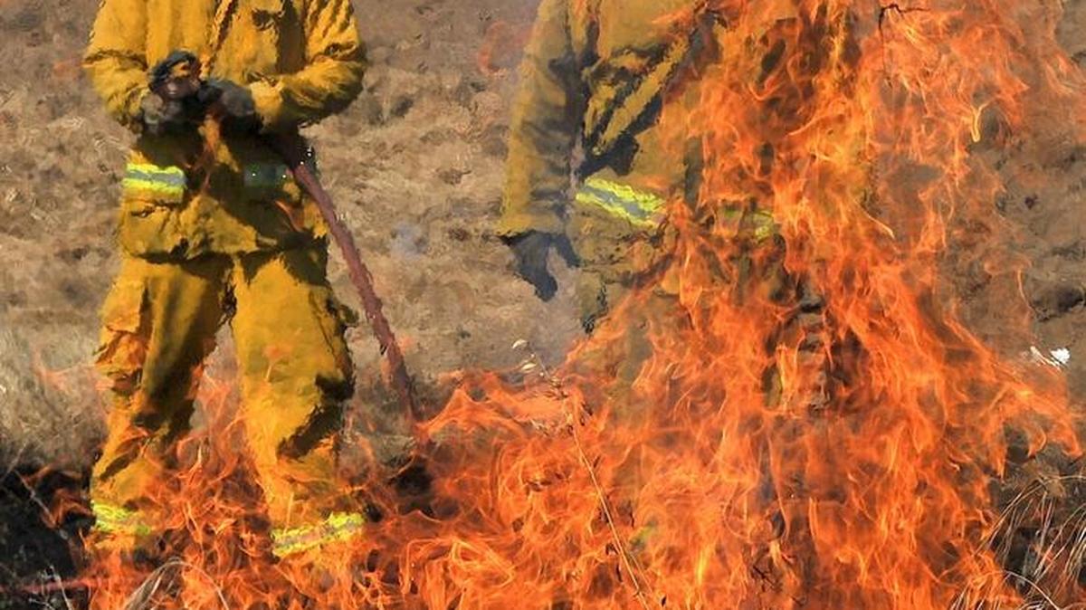 Fresno firefighter Phillip Souza, left, and Capt. Pete Martinez are seen through heat waves as they watch a fire quickly ignite from a legal fountain-type firework during a demonstration of the effects of fireworks around dry grass areas on Tuesday, June 28, 2016.