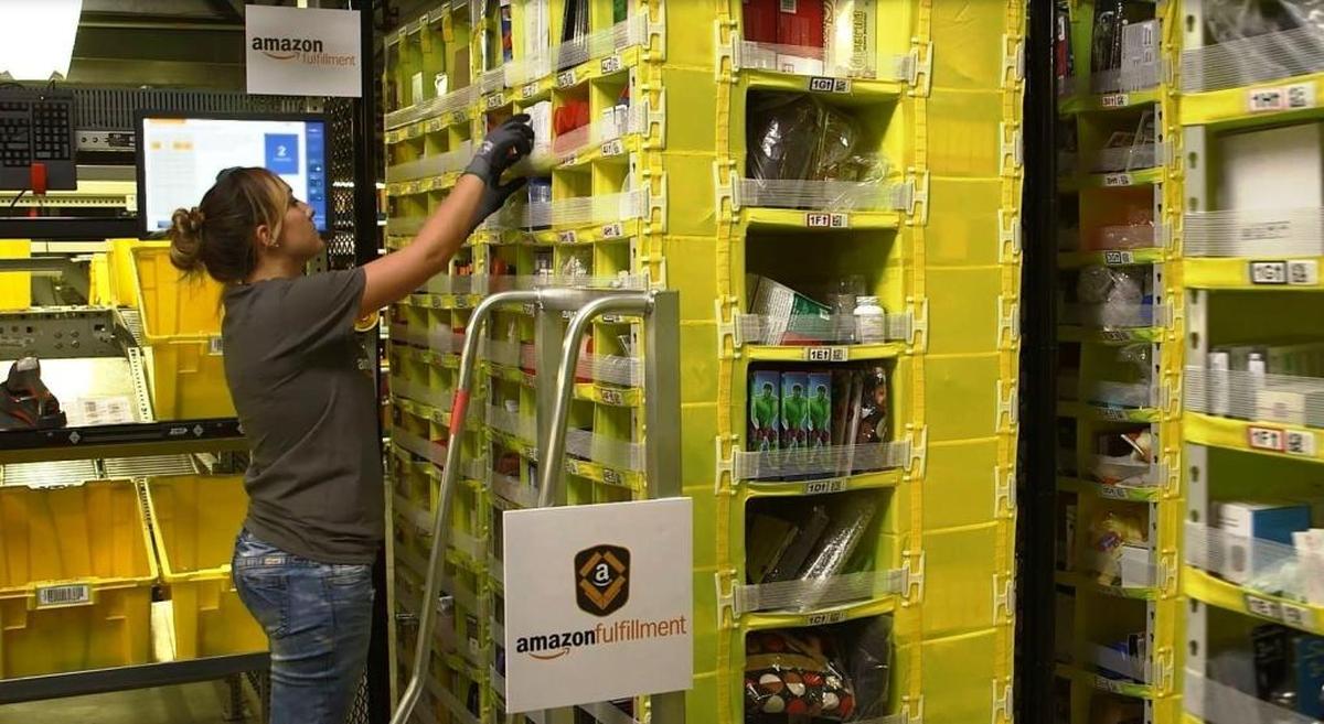 A worker in an Amazon fulfillment center grabs a product from a bin as she assembles a customer’s order for shipping in this image captured from a company video. The centers use automated robots to ferry racks of product bins to the workers.
