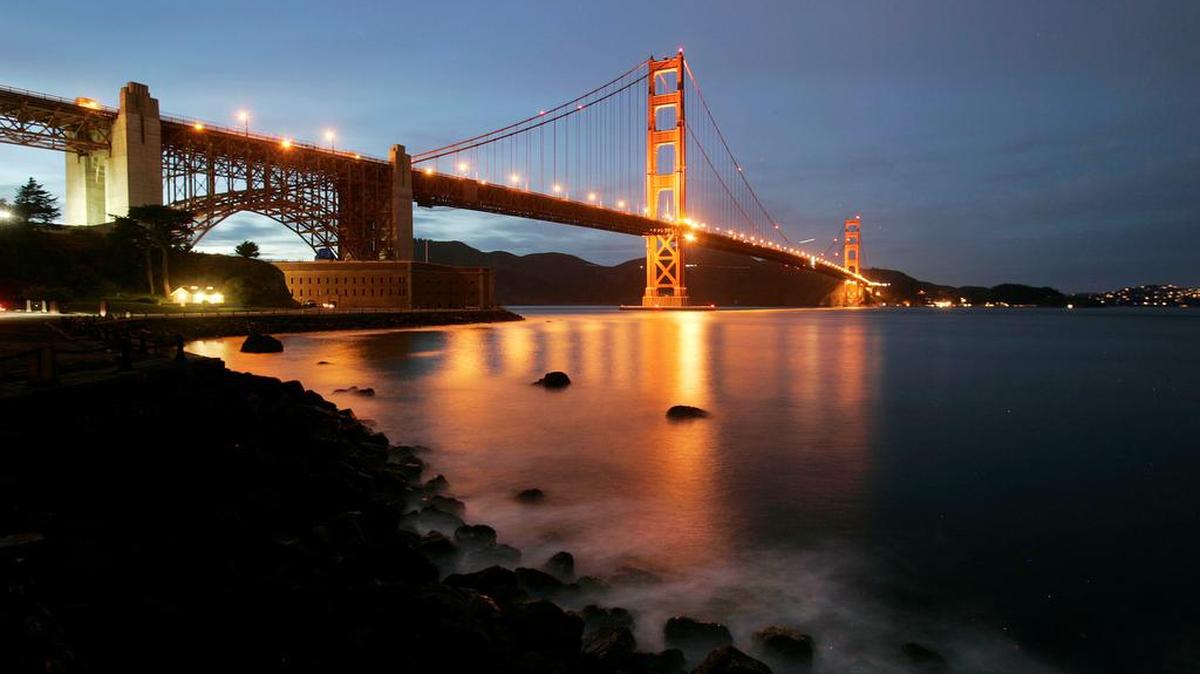 Waves crash on the pier at Fort Point as the Golden Gate Bridge lights up in the background at dusk in San Francisco.