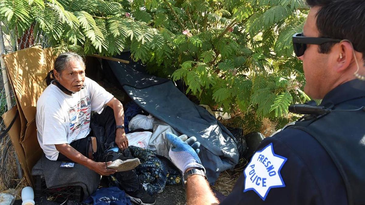 A homeless man puts his shoes on as Fresno police officer Noel Perez offers assistance after telling the man he needs to leave his camp, and take what he can carry, Wednesday morning, Aug. 14, 2018.