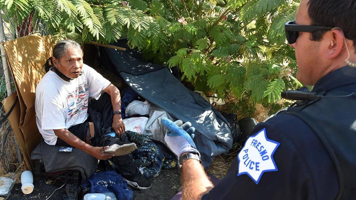 A homeless man gets his shoes on as Fresno police officer Noel Perez tells the man he needs to leave his camp, nearly shrouded by a tree, and take what he can carry, Wednesday morning, Aug. 14, 2018.