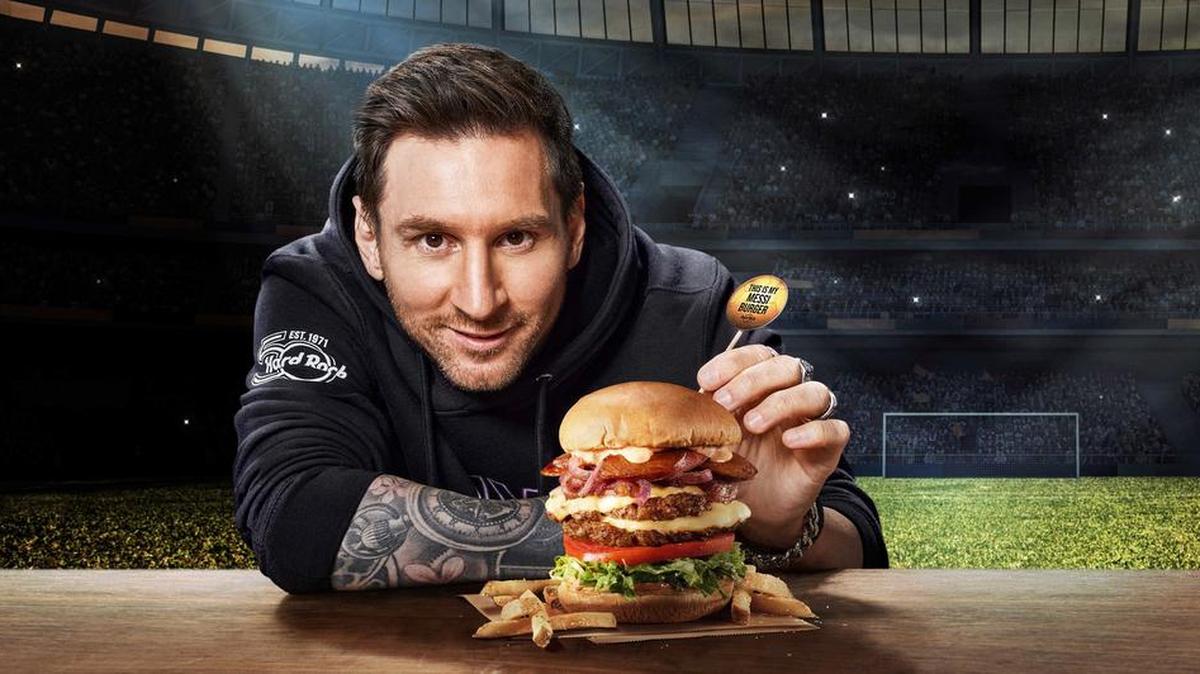 Fotografía promocional cedida por Hard Rock International donde aparece el jugador argentino de fútbol Leo Messi mientras muestra una enorme hamburguesa bautizada ‘Messi Burger’ presentada en un pan brioche tostado con dos porciones de carne, además de queso provolone, chorizo en rodajas, cebolla roja caramelizada, lechuga romana y rodajas de tomate, todo ello aderezado con una salsa picante y ahumada.