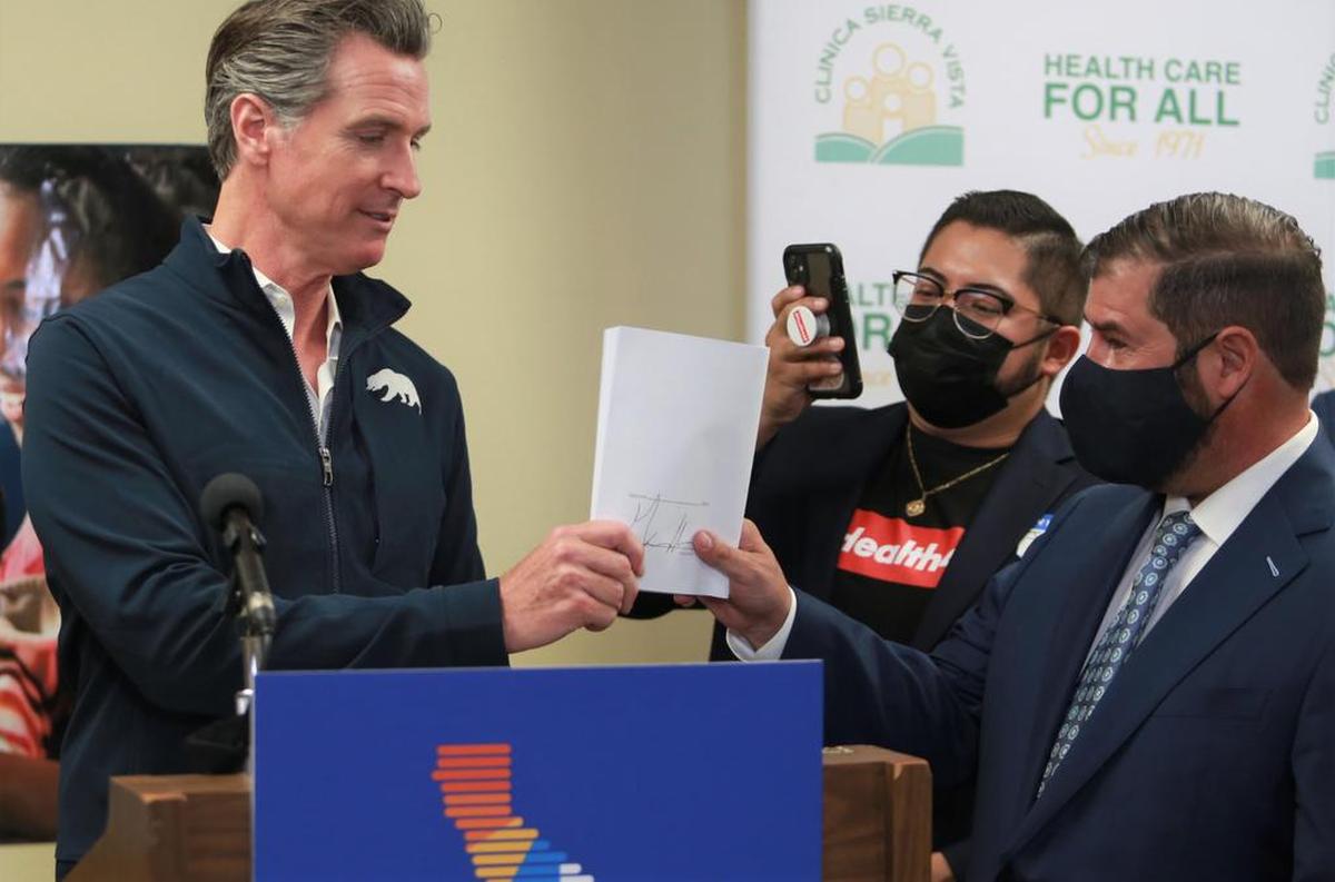 El gobernador de California, Gavin Newsom, izquierda, entrega la documentación al Asambleísta Joaquín Arambula, D-Fresno, después de firmar el proyecto de ley que amplía la cobertura de atención médica para los californianos indocumentados en la Clínica Sierra Vista el martes 27 de julio de 2021 por la mañana en Fresno, CA.